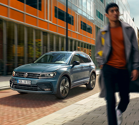 Ein blauer VW T-Roc fährt eine Straße in einer Großstadt entlang. Im Vordergrund geht ein junger Mann auf einem Bürgersteig.