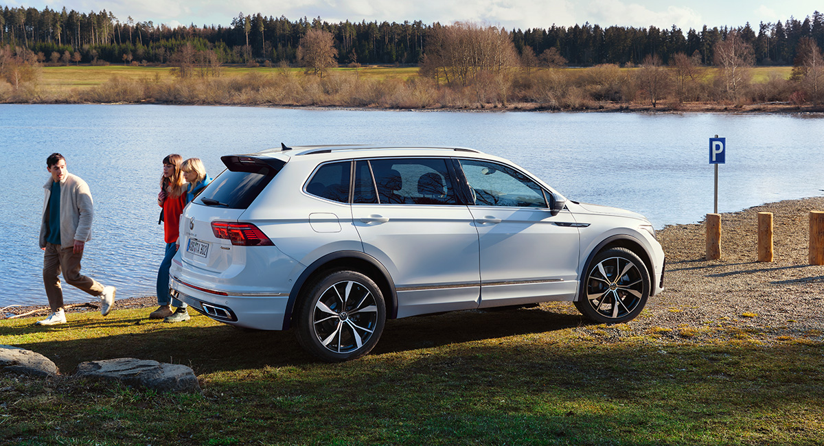 Weißer VW Tiguan Allspace von der Seite vor einem See. Links gehen drei Menschen entlang.
