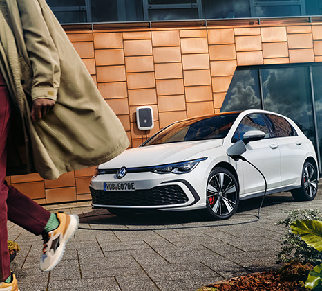 Ein weißer VW Golf GTE ist vor einem modernen Haus geparkt. In der Ladeklappe steckt das Ladekabel, das an eine ID. Charger Wallbox angeschlossen ist. Am linken Seitenrand geht eine Person aus dem Bild.