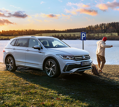 Weißer VW Tiguan Allspace im Dreviertelprofil vor einem See. Im Hintergrund lässt ein Mann Steine über die Wasseroberfläche hüpfen.